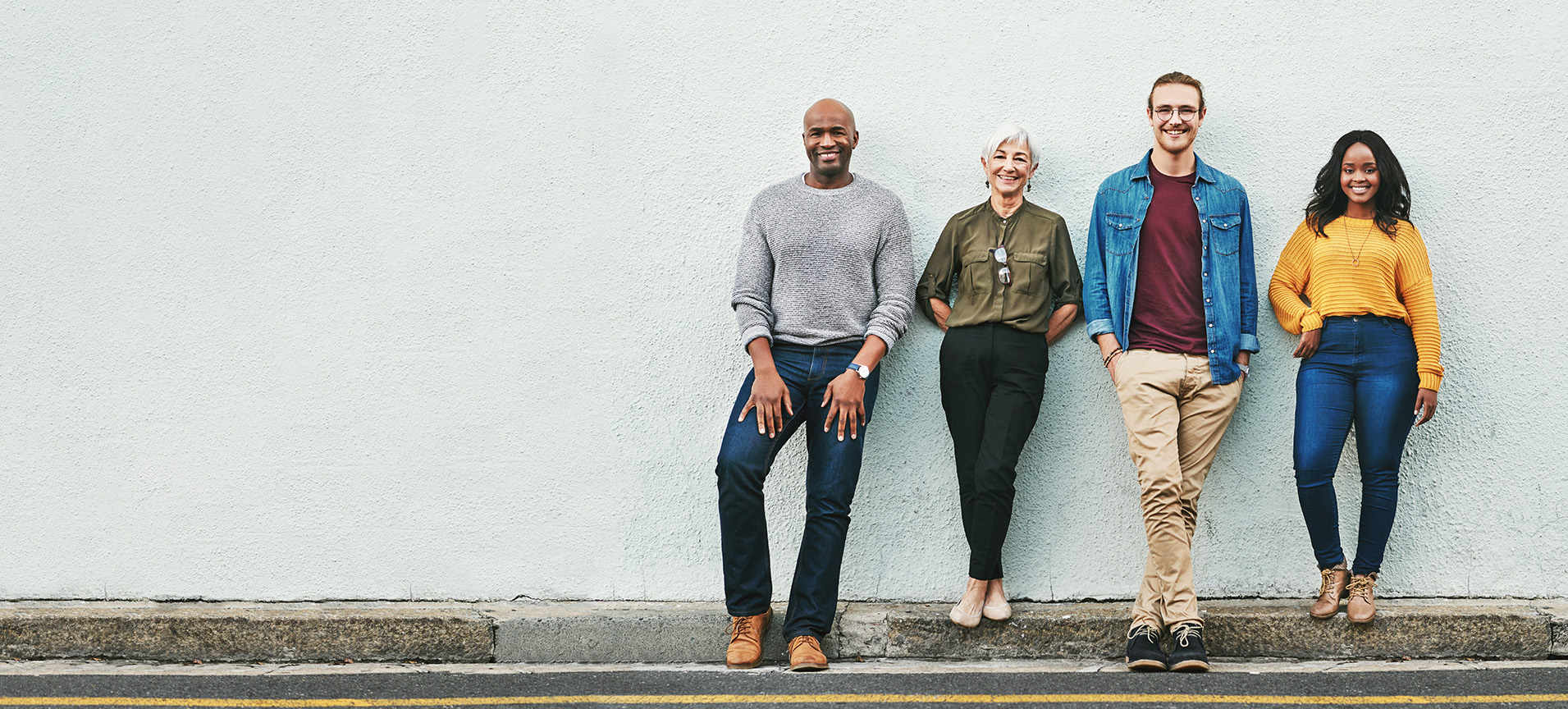 Four smiling adults lean on a white wall outside