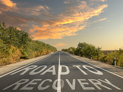 road to recovery is written on a highway that is on a scenic route through trees, leading to a sky with pretty clouds